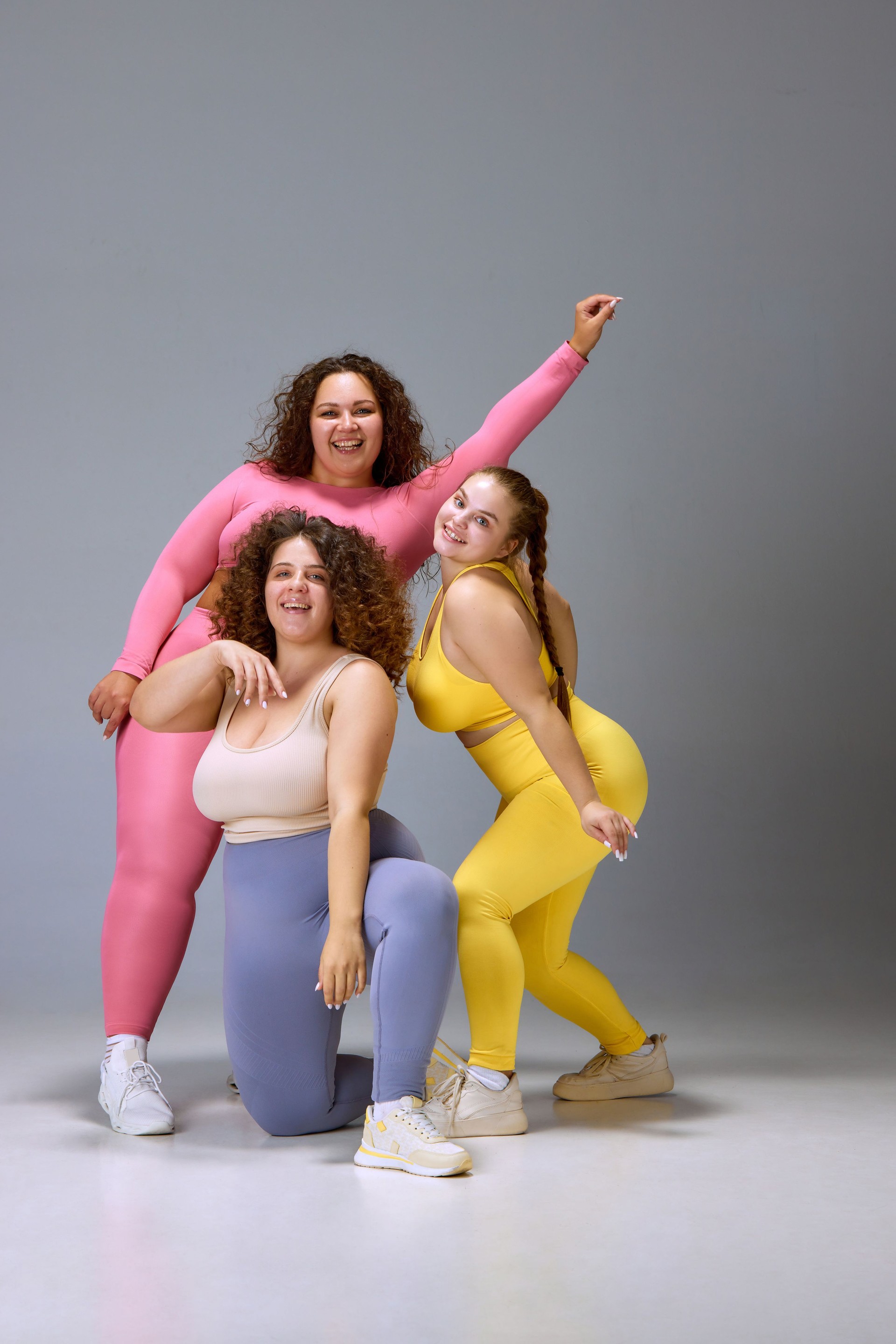 Acceptance. Three smiling young women in comfortable sportswear standing against grey studio background. Plus size beauty