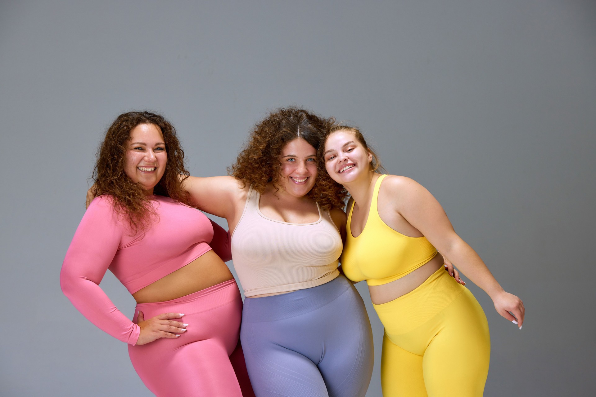 Support. Three beautiful women with oversized bodies, wearing sportswear, posing against grey studio background